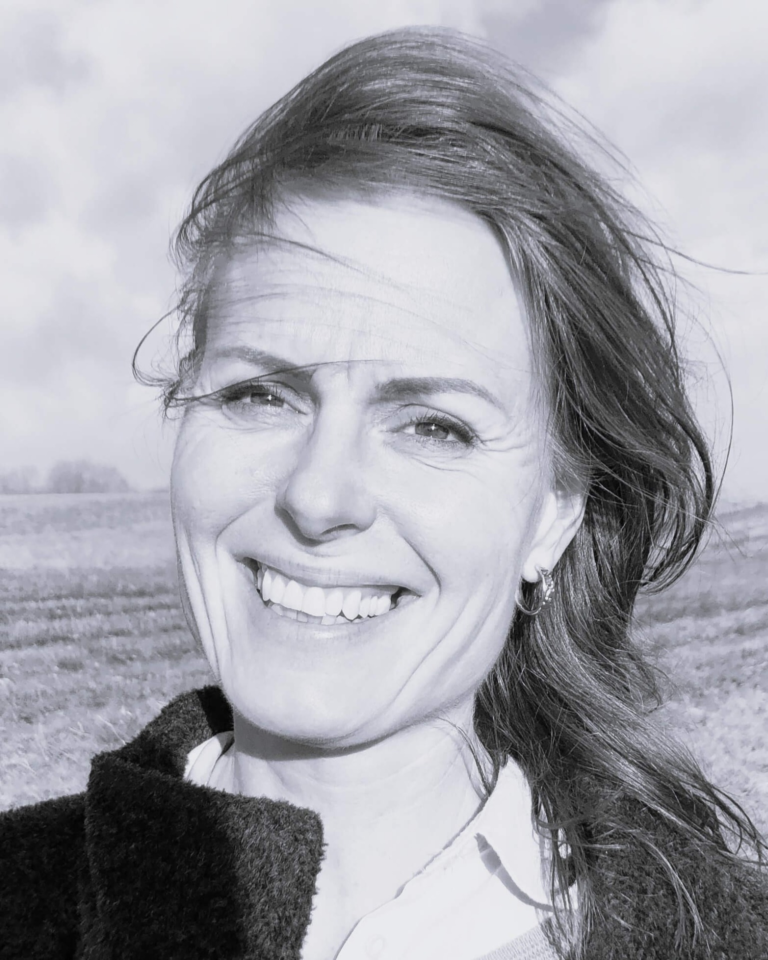 Sort/hvid nærbillede af smilende kvinde med vindblæst hår og øreringe, udendørs. Anette Werenskiold